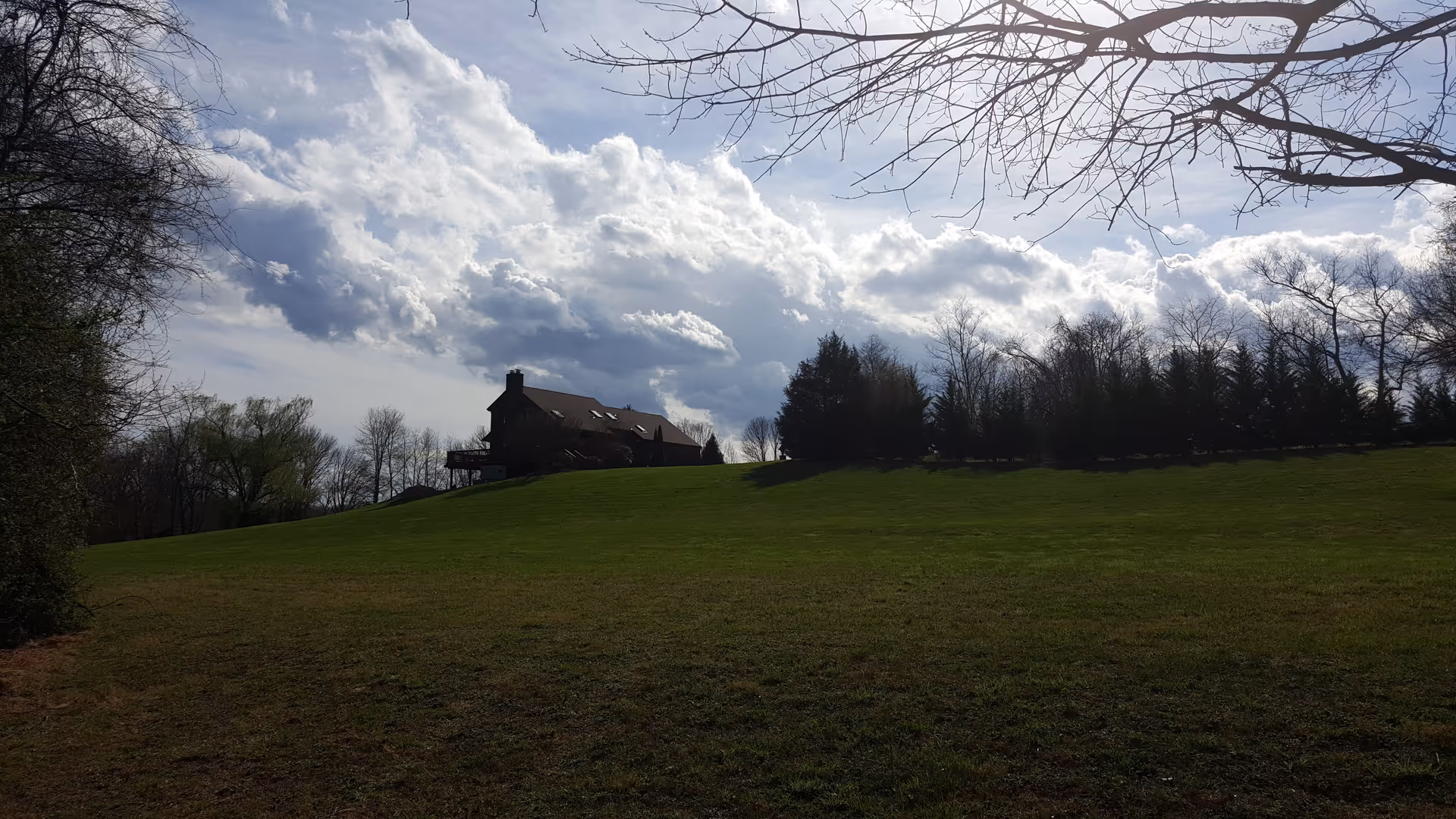 A grassy hill with a brown building at the crest, trees and cloudy sky backdrop.