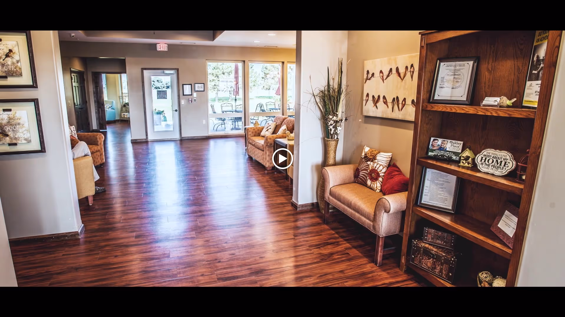 Spacious senior living lobby with wooden floors, armchairs, decorative shelving, and glass doors opening to an outdoor patio.
