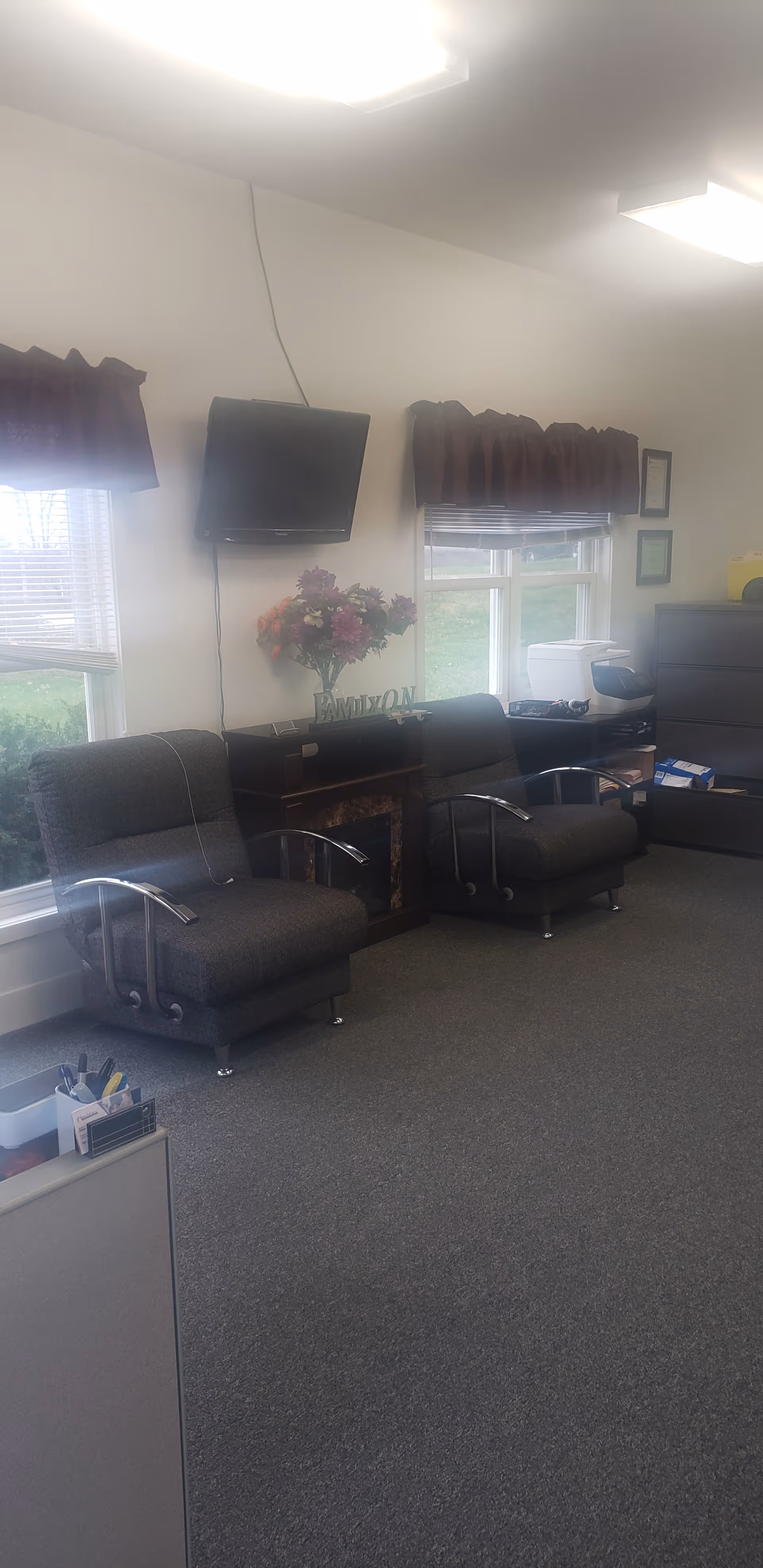A quiet lounge area with two dark upholstered chairs, a wall-mounted TV, and a flower arrangement.
