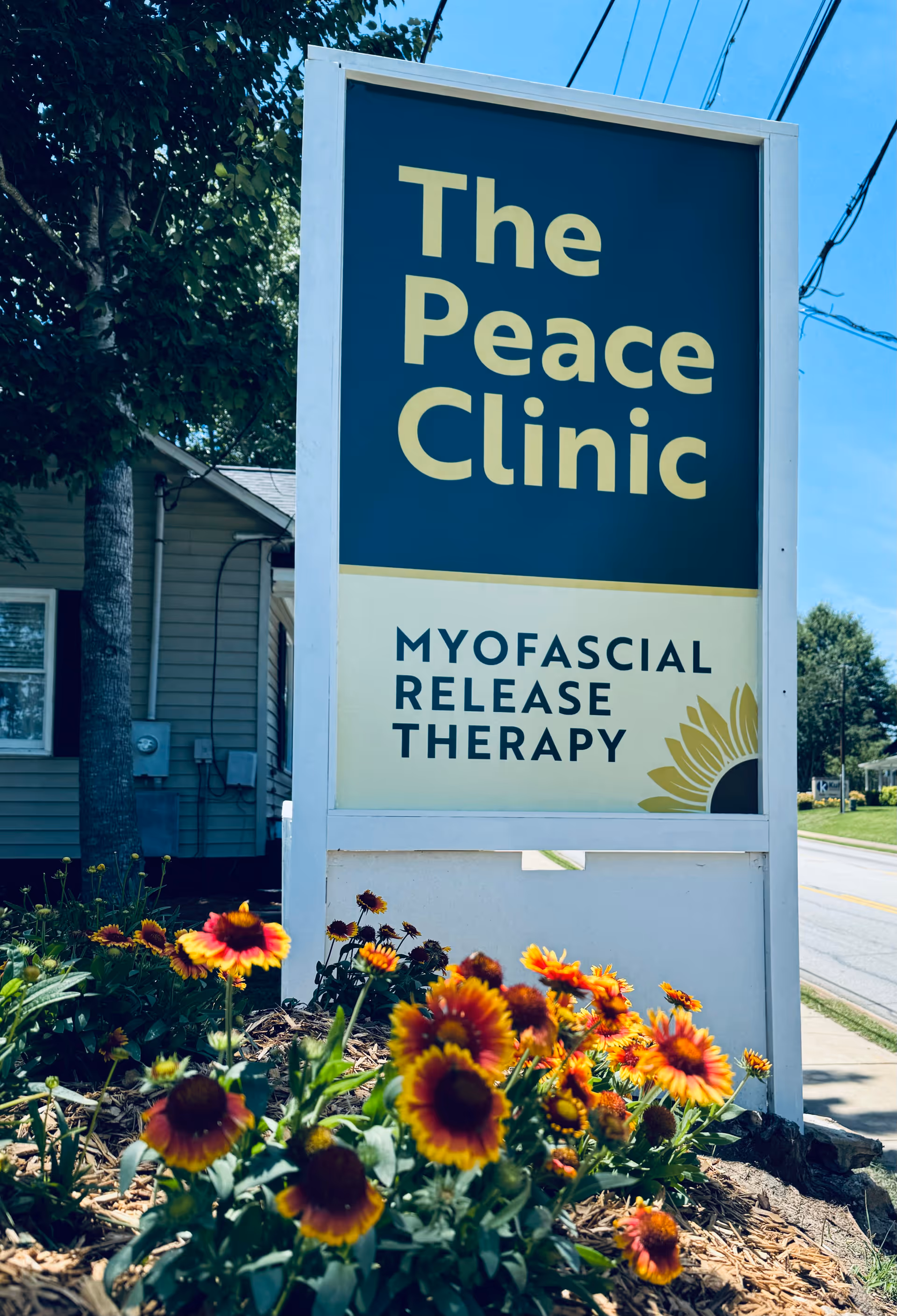 Outdoor sign for The Peace Clinic, Myofascial Release Therapy, with colorful flowers in foreground