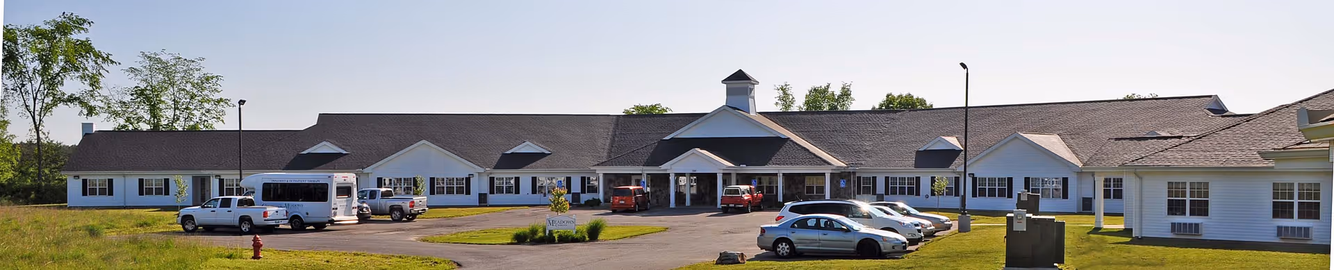 Front exterior of a single-story senior living facility with a covered entrance, parked cars, and surrounding lawn.