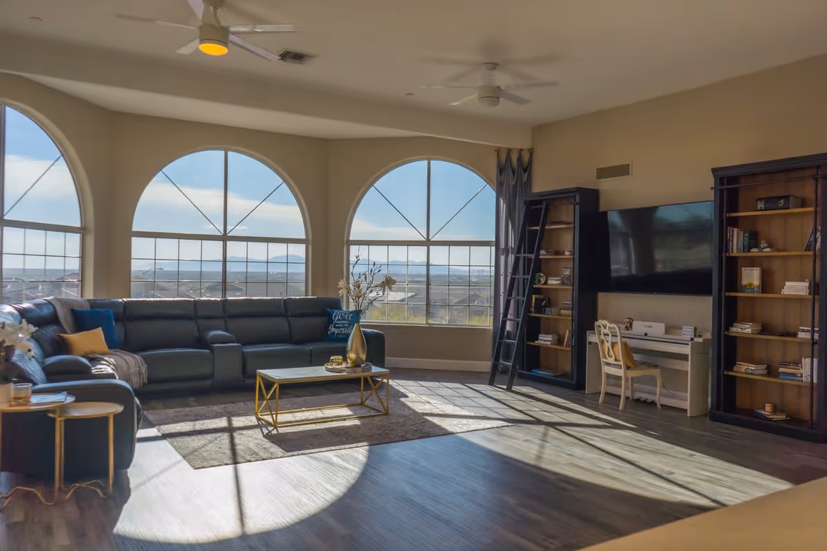 A spacious living room with large arched windows letting in natural light. The room features a dark sectional sofa with pillows, a coffee table with a vase of flowers, two tall bookshelves with a ladder, a wall-mounted flat-screen TV, and a white desk with a chair. The floor is wooden, and ceiling fans are visible.
