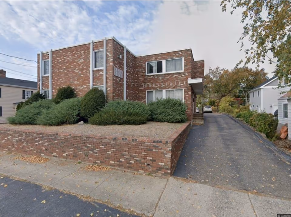 Brick senior living facility front view with driveway, shrubs, and a parked car.