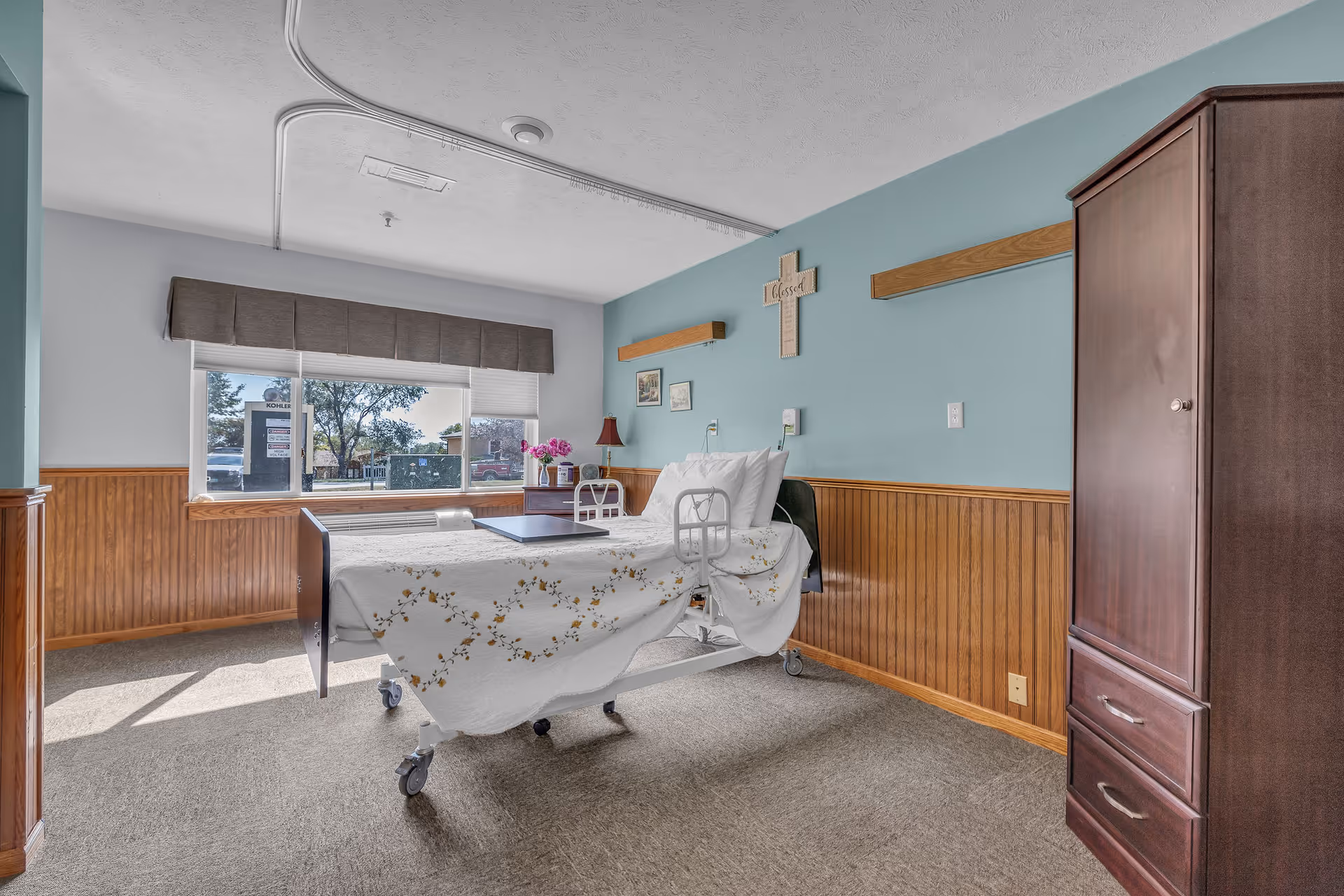 A bright and clean patient room in a healthcare facility with a hospital bed covered in a white floral bedspread, a wooden wardrobe, a window with a view of trees and parked cars, and light blue walls with wooden paneling halfway up. A cross with the word 'Blessed' hangs on the wall above the bed.