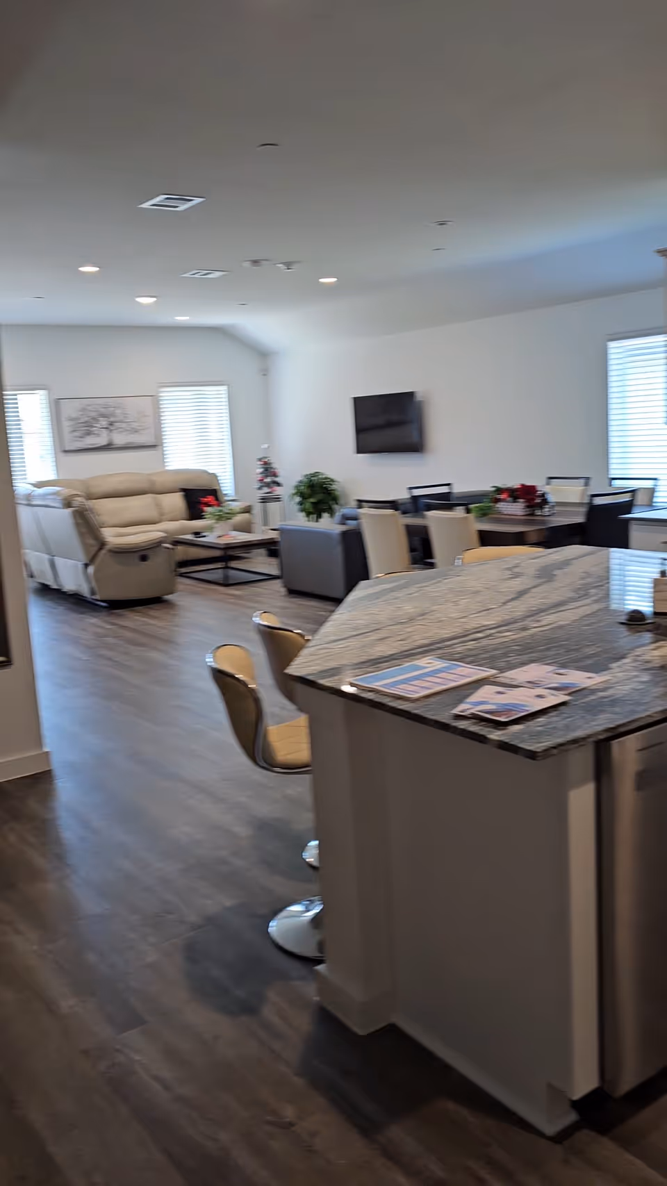 Open lounge with a marble kitchen island, seating, TV, and windows in a senior living space.