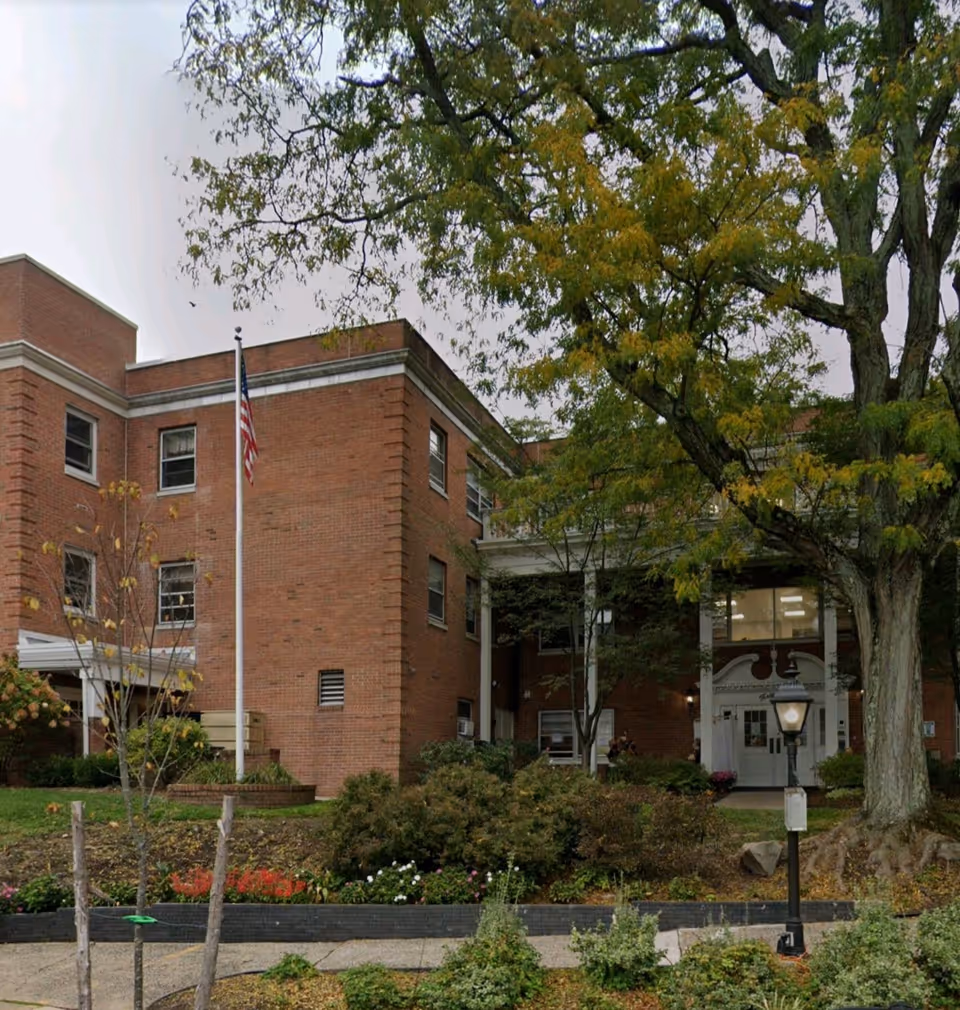 Brick senior living facility exterior with flag, trees, and entrance.