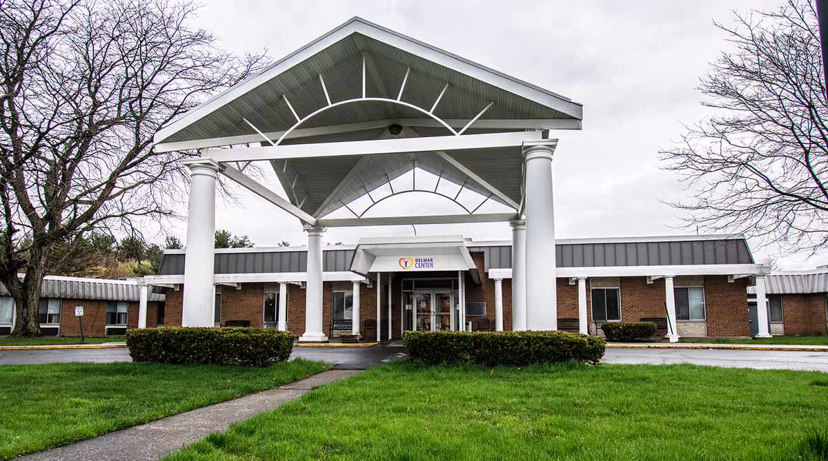 Front exterior view of the Delmar Center for Rehabilitation and Nursing building with a large white covered entrance supported by columns, surrounded by green grass and leafless trees under a cloudy sky.