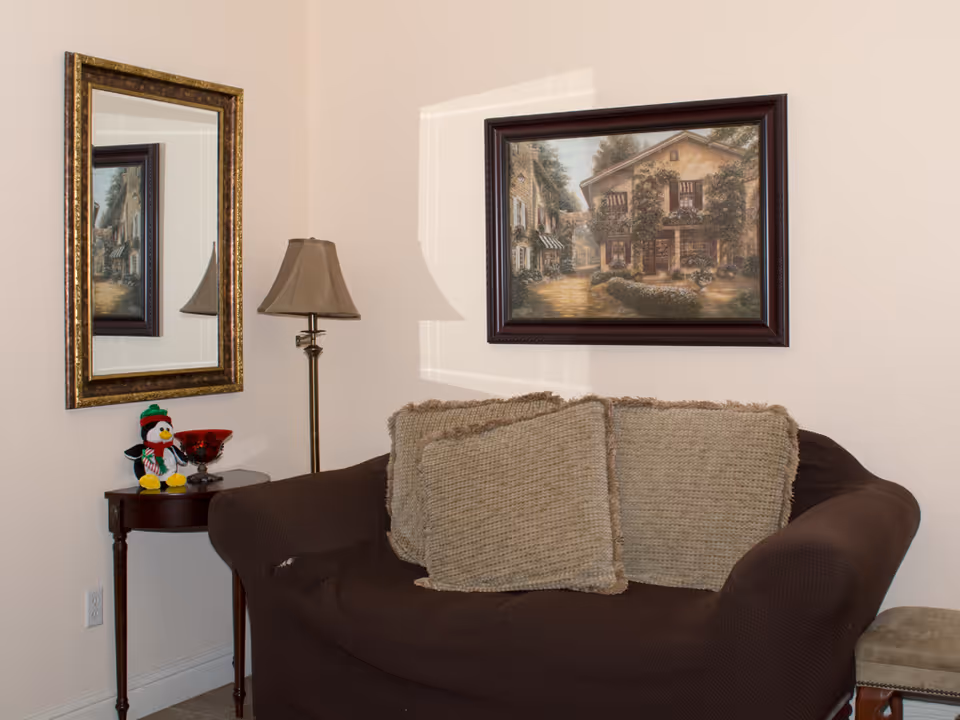 A cozy corner of a living room with a dark brown loveseat covered with two large beige cushions. Above the loveseat hangs a framed painting of a rustic house with greenery. To the left of the loveseat is a small wooden side table with a decorative red bowl and a small stuffed penguin toy wearing a green hat and scarf. A tall floor lamp with a beige lampshade stands behind the table. A large rectangular mirror with a gold frame is mounted on the adjacent wall, reflecting part of the painting.