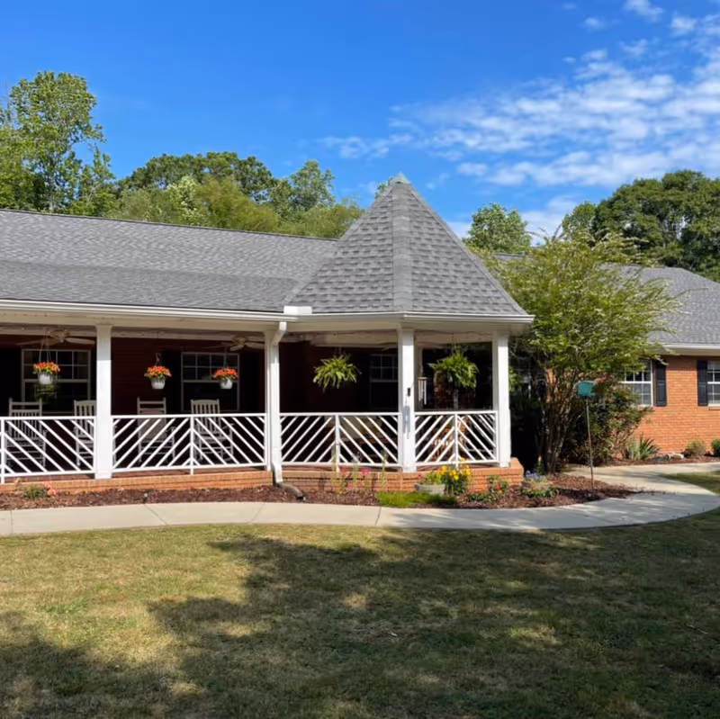 Exterior view of a brick building with a covered porch featuring white railings and hanging flower pots. The porch has ceiling fans and is surrounded by a well-maintained lawn and trees under a blue sky with some clouds.