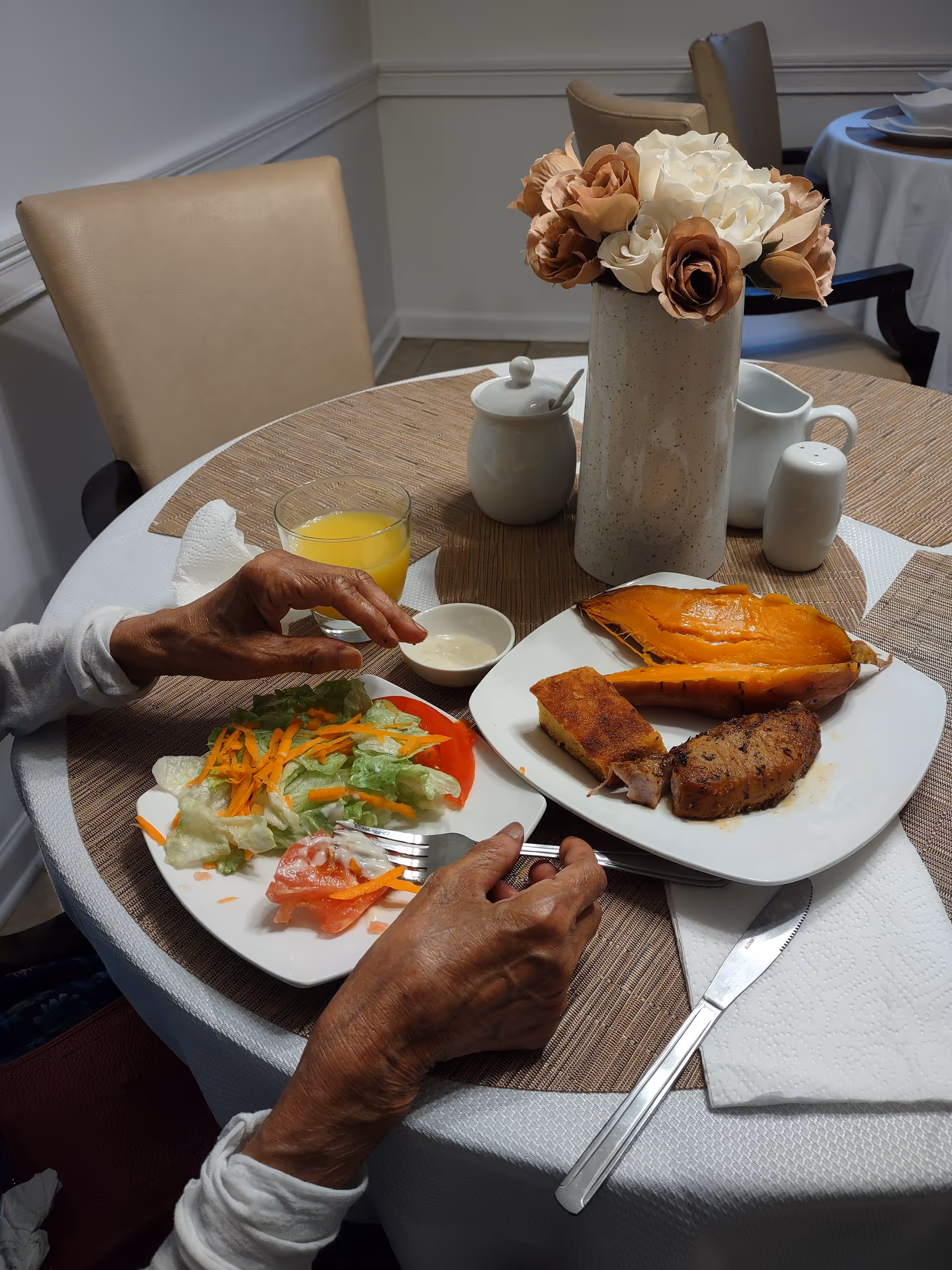 Elderly hands dine at a round table with salad, meat, juice, and a floral centerpiece.