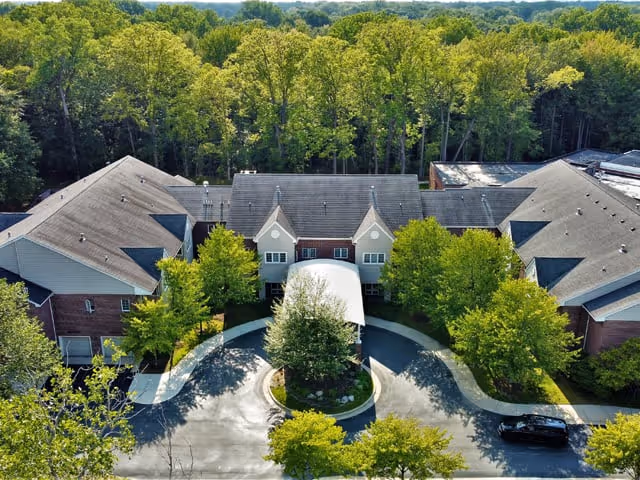 Aerial view of a senior living facility entrance with a covered drop-off, circular driveway, surrounding trees, and adjacent buildings.