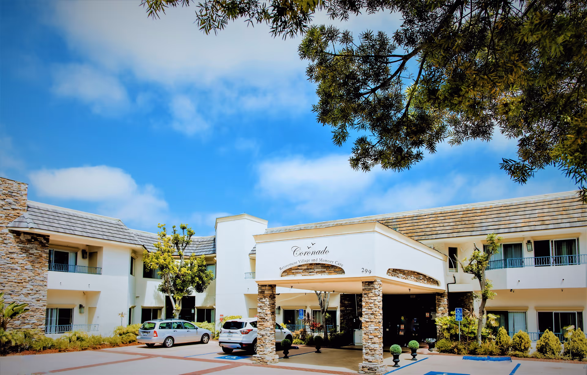 Exterior view of Coronado Retirement Village entrance with stone pillars, parked cars, and blue sky.