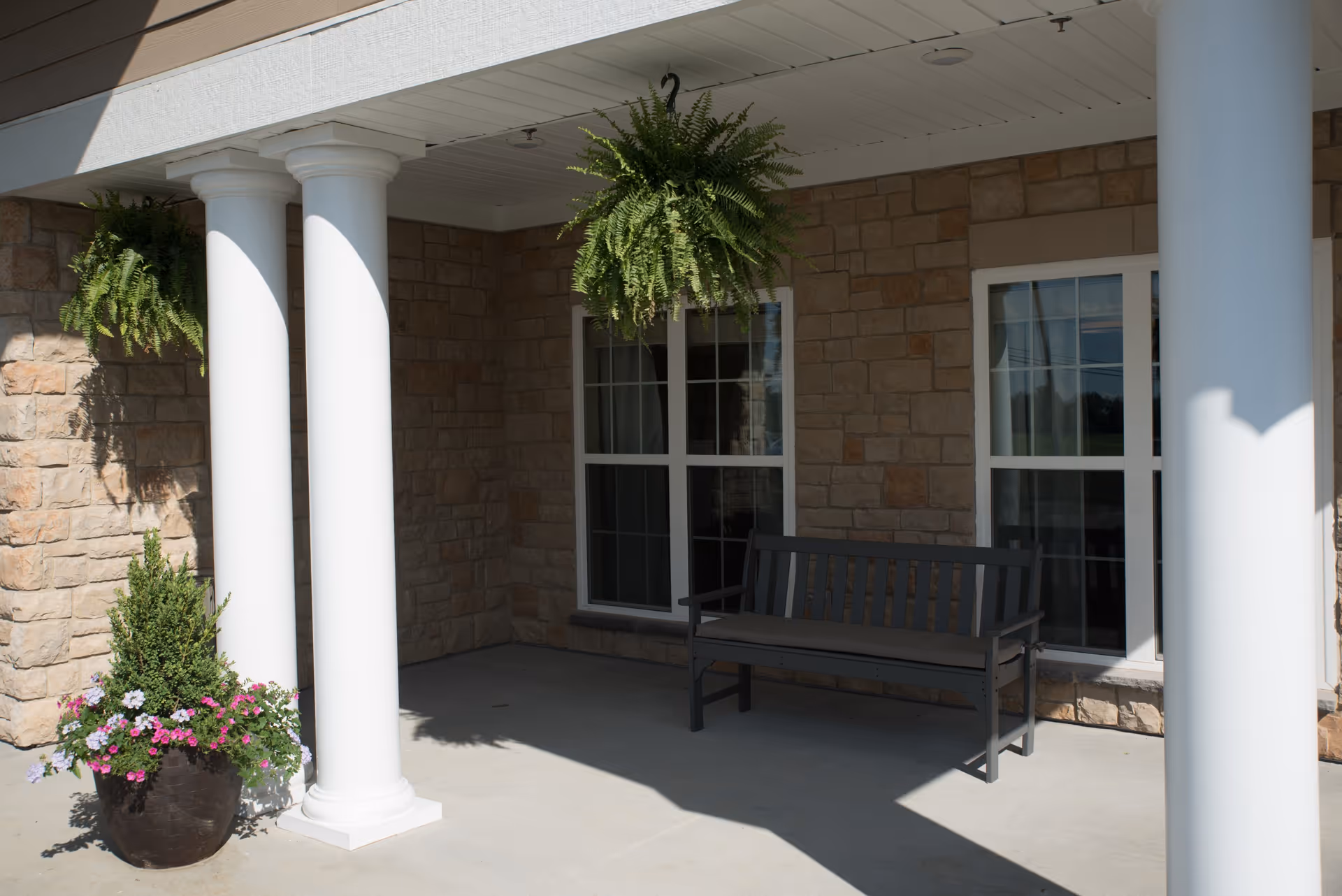 Covered front porch with white columns, a bench, hanging ferns, and a potted flower arrangement against a stone exterior.