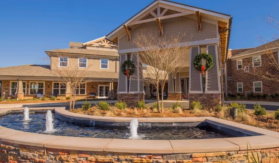 Front entrance of a senior living facility with a circular fountain, stone facade, and holiday wreaths on the porch.