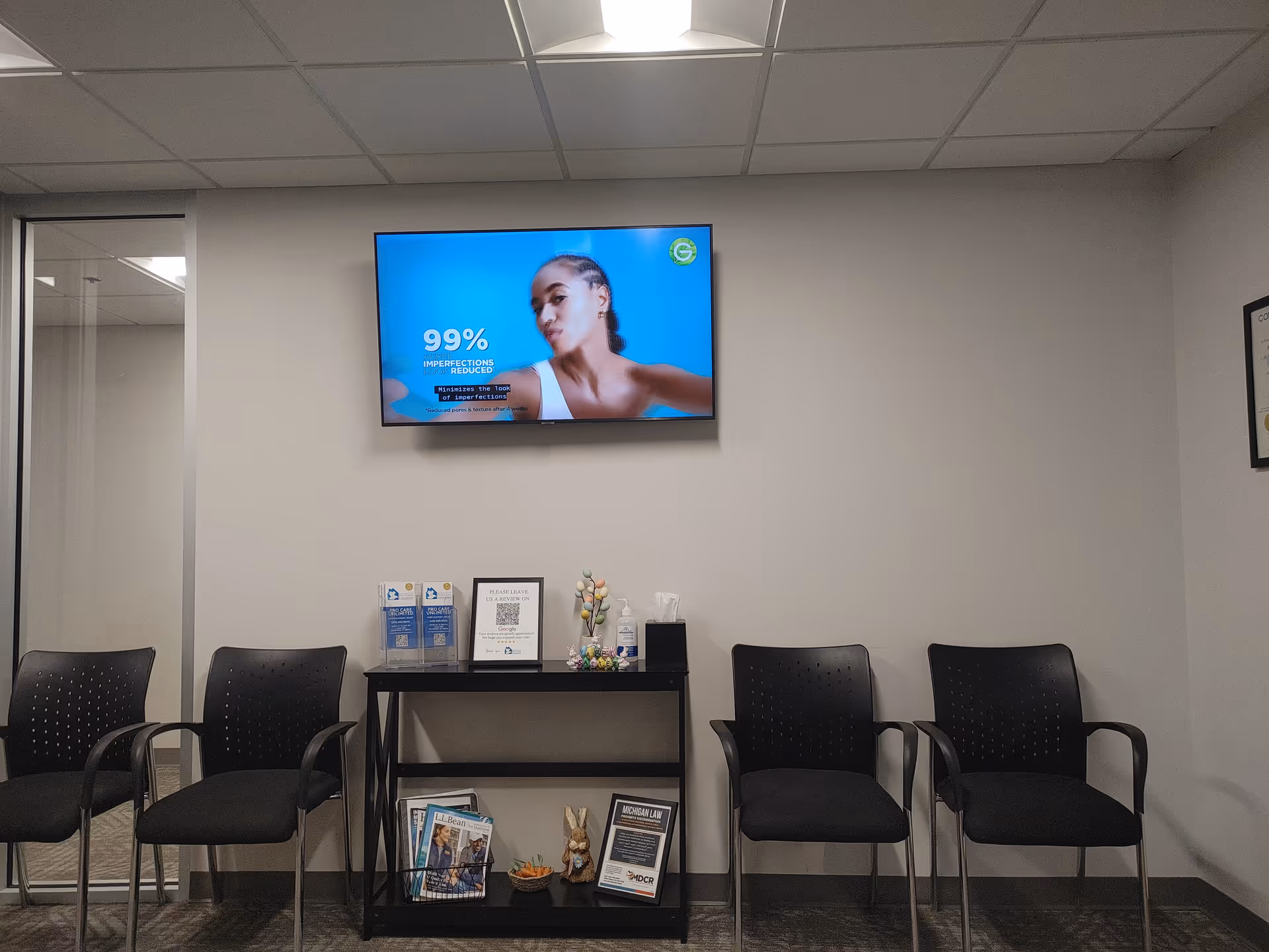 Indoor waiting area with black chairs, a wall-mounted TV, and a small display table.