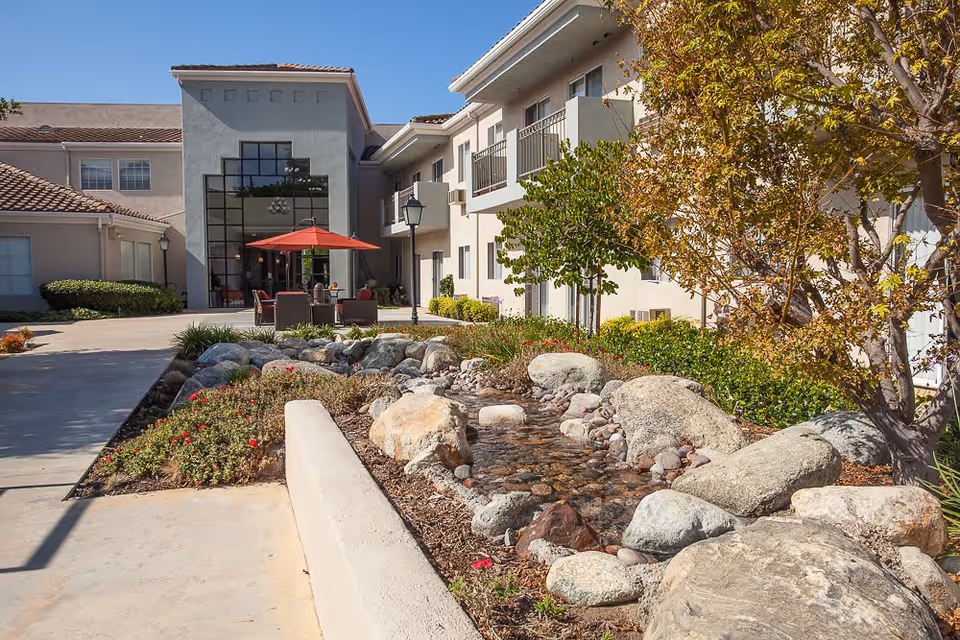 Outdoor view of Walnut Valley Senior Living facility showing a landscaped garden with rocks and a small water feature in the foreground. The building has large windows and balconies, with a patio area featuring a red umbrella and outdoor seating.