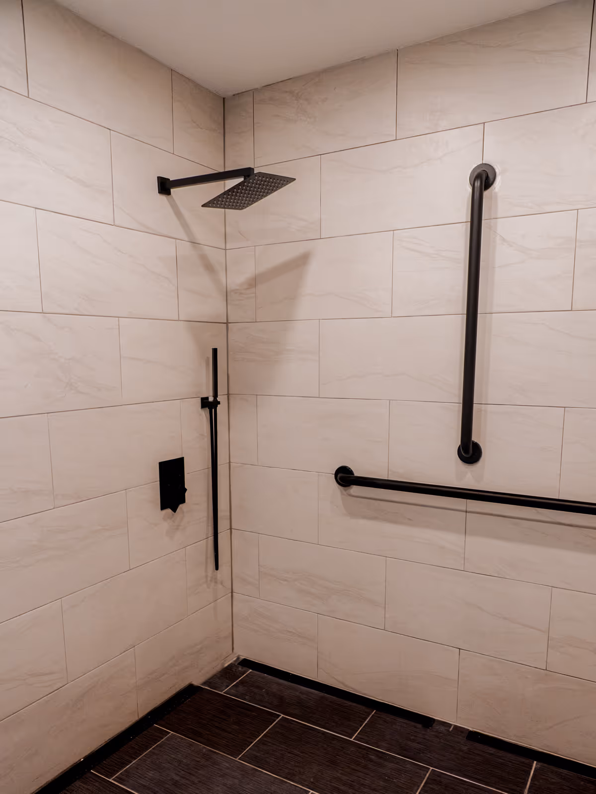 Accessible tiled shower with a rainfall head, black grab bars, and dark floor tiles.