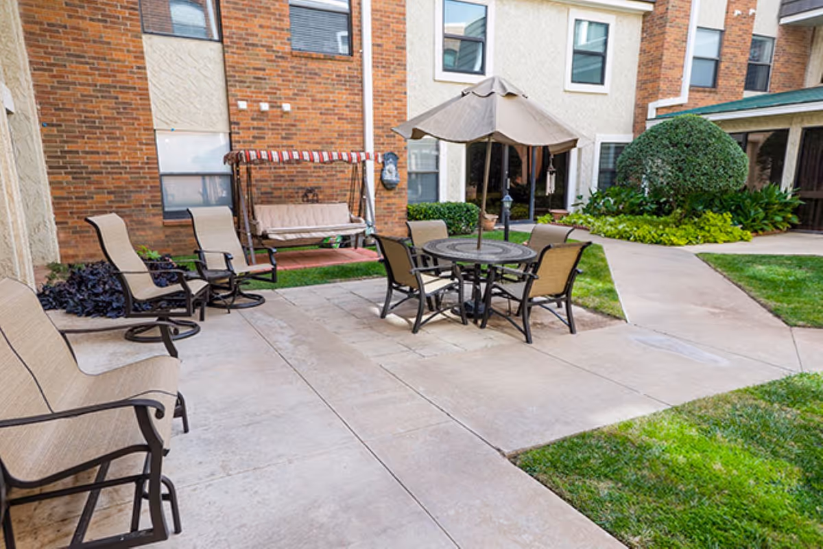 Outdoor patio at Brookdale Grand Court Lubbock with seating, umbrella, swing bench, and greenery.