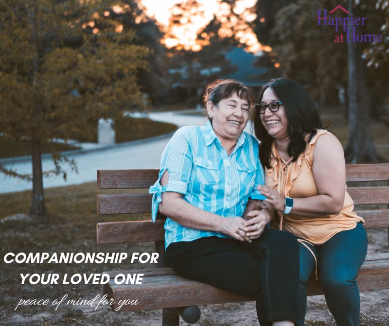 Two women sit on a wooden outdoor bench, smiling, with Happier at Home branding.