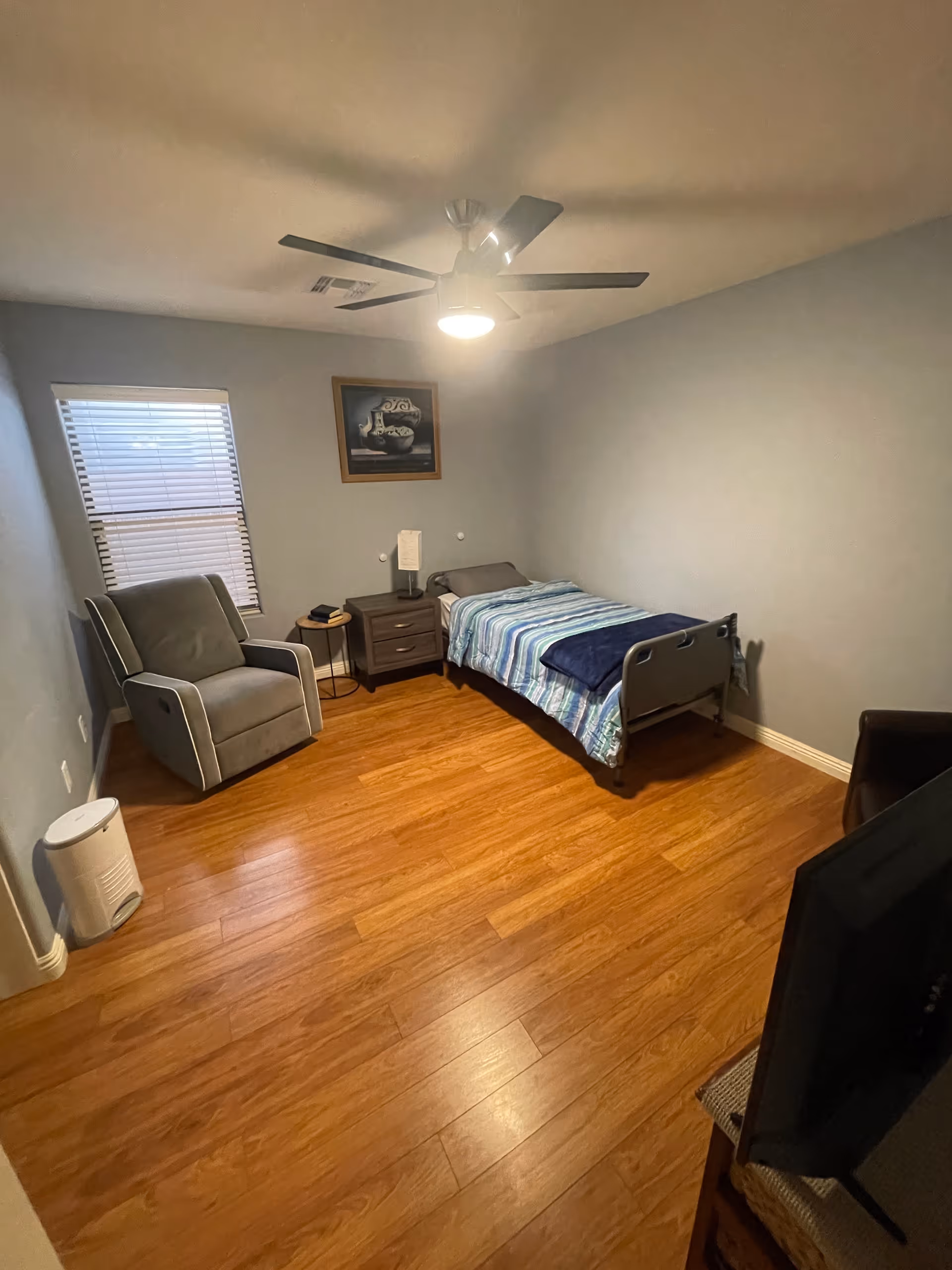 Neatly furnished bedroom with a single bed, chair, nightstand, window blinds, and a ceiling fan.