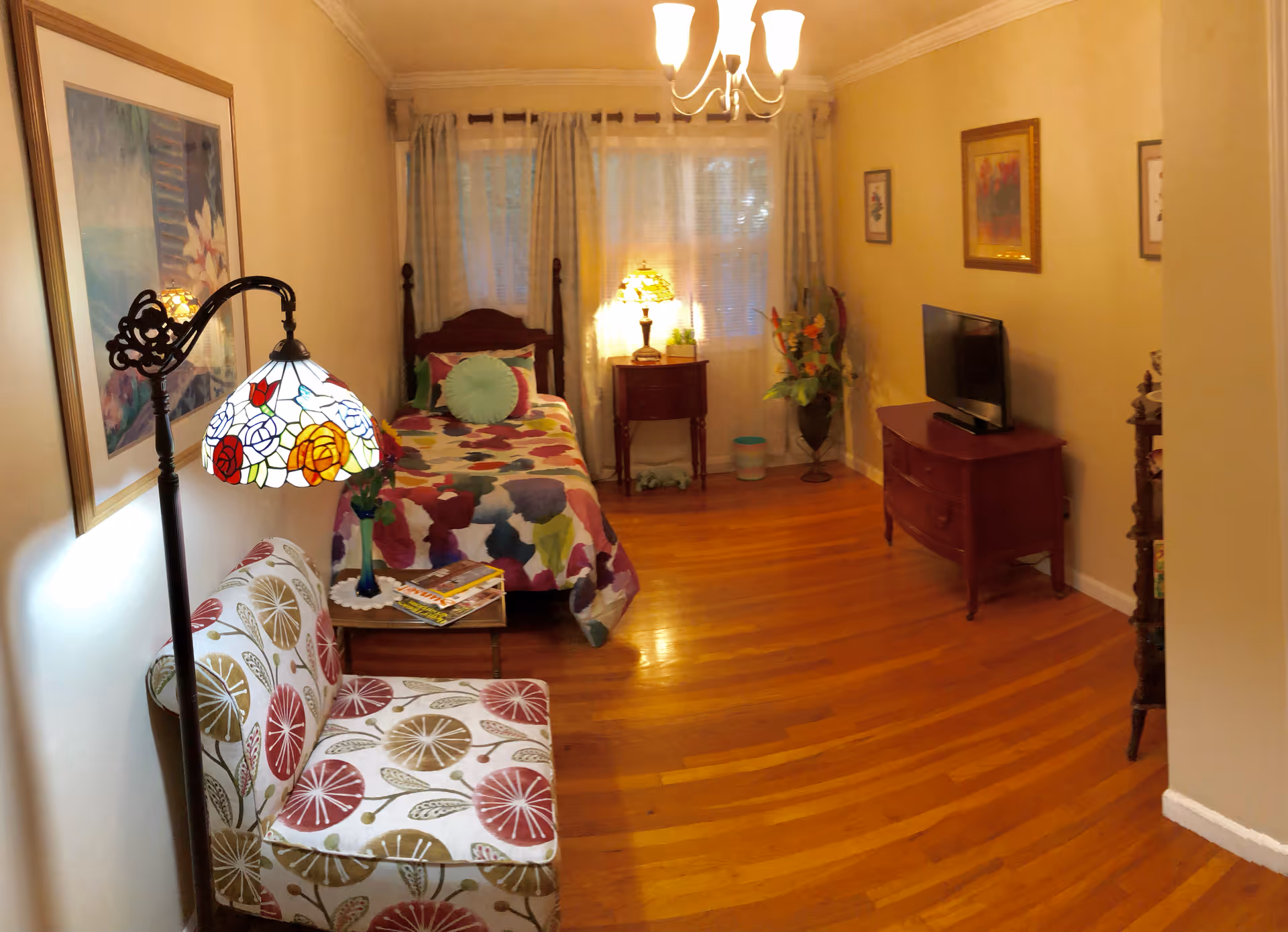 A cozy bedroom with a single bed covered in a colorful floral quilt, a small wooden nightstand with a lamp, a patterned armchair, a floor lamp with a stained glass shade, a wooden dresser with a TV on top, and framed artwork on the walls. The room has hardwood floors and light-colored walls with a window covered by curtains.