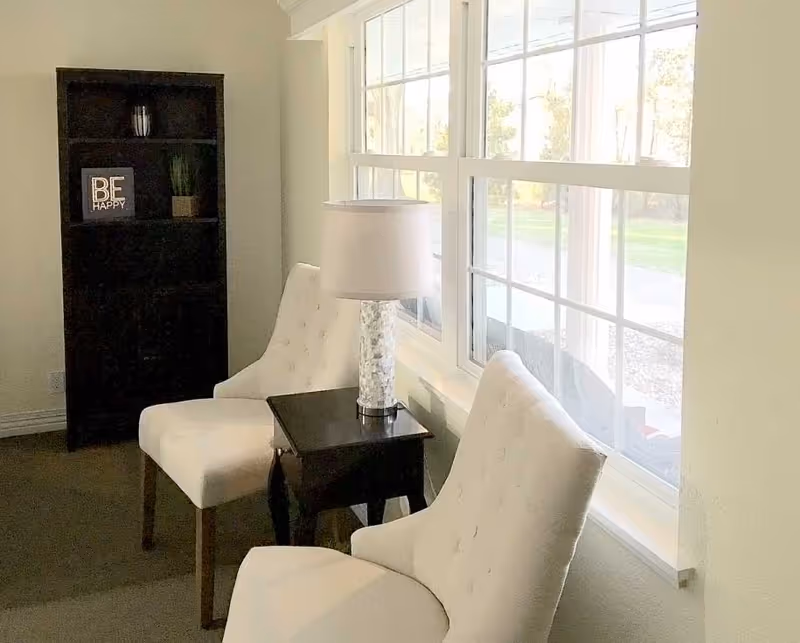 A cozy sitting area with two white upholstered chairs positioned around a small dark wooden side table with a decorative lamp. Behind the chairs is a tall dark wooden bookshelf with a small plant and a framed sign that says 'BE HAPPY'. Large windows let in natural light and offer a view of greenery outside.