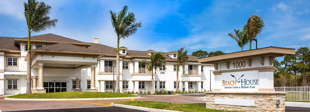 Exterior view of the Beach House Assisted Living & Memory Care facility with a large entrance, palm trees, and a sign displaying the facility name and address number 1000.