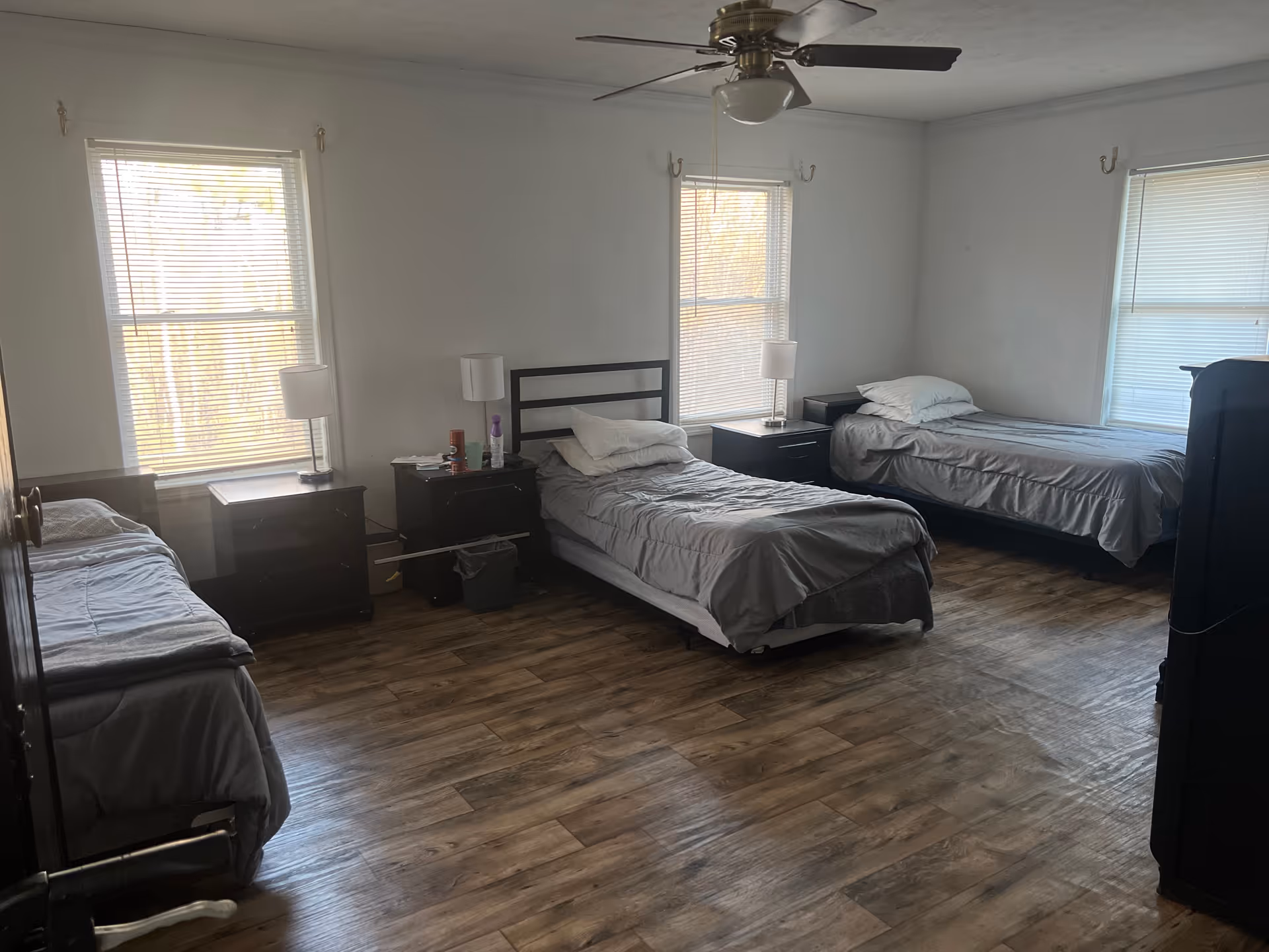 A shared bedroom with three single beds, nightstands, lamps, ceiling fan, and windows.