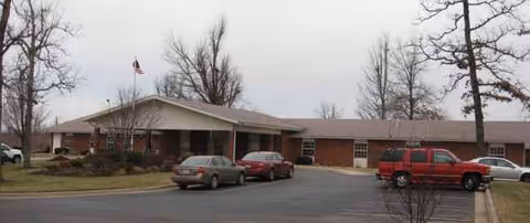 Single-story brick senior living facility with a front entrance, parked cars, and an American flag.