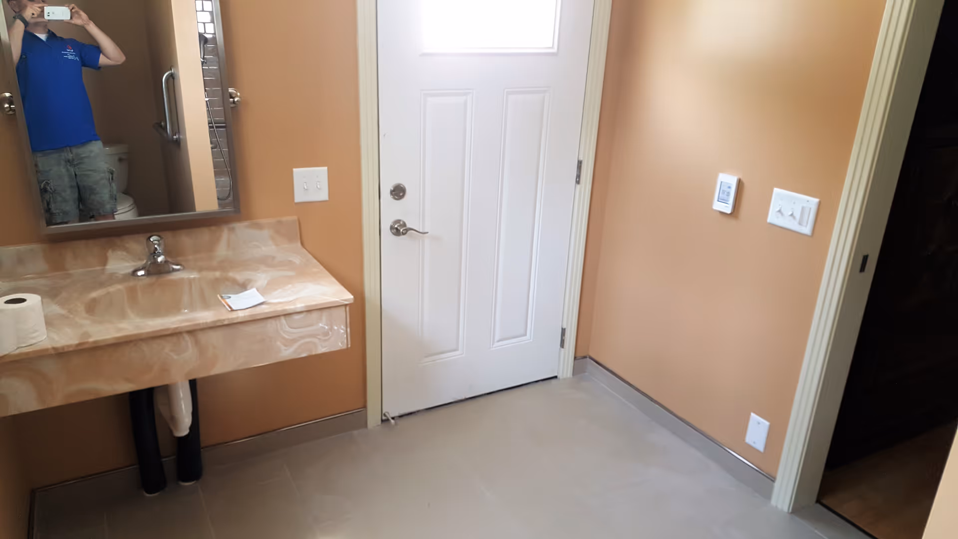 Peach colored bathroom with sink, mirror, door, and a person visible in the mirror.