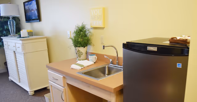 A small kitchenette area with a stainless steel sink, a potted plant, a cup, and a folded towel on the countertop. To the right is a compact black refrigerator. In the background, there is a white cabinet with drawers and a lamp on top, and a wall-mounted TV above it. A yellow wall decoration reads 'YOU ARE MY SUNSHINE'.