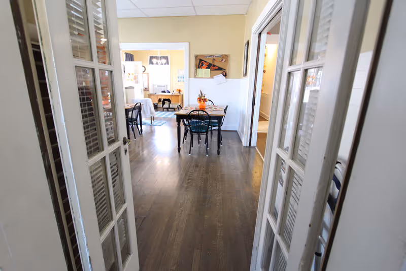 Bright dining room inside a senior living facility with tables, chairs, and double doors.