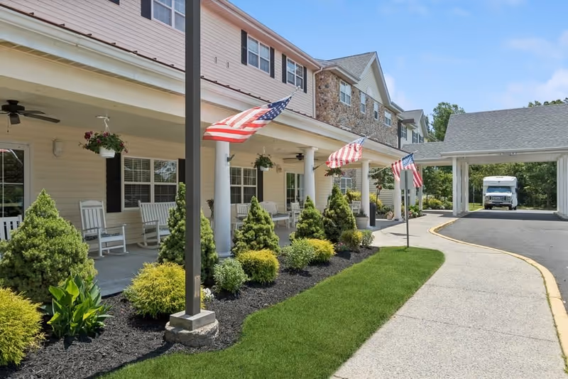 Exterior view of Washington Township Senior Living facility showing a covered porch with white rocking chairs, hanging flower pots, American flags, and well-maintained landscaping with shrubs and green grass. A driveway curves alongside the building leading to a covered entrance with a vehicle parked underneath.
