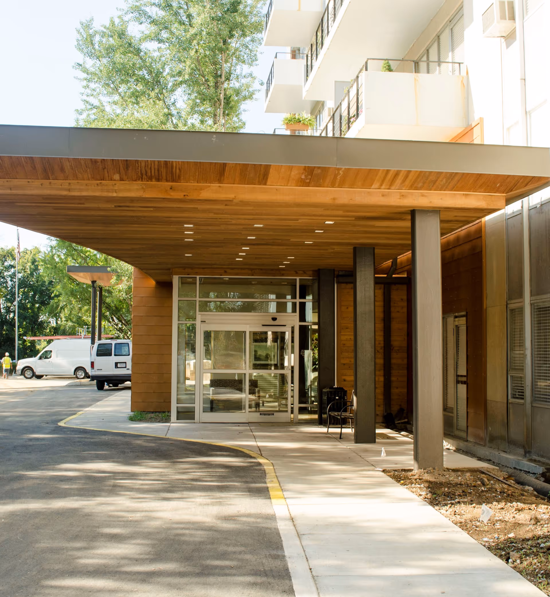 Modern building entrance with a wooden canopy, glass doors, and parked white vans outside.