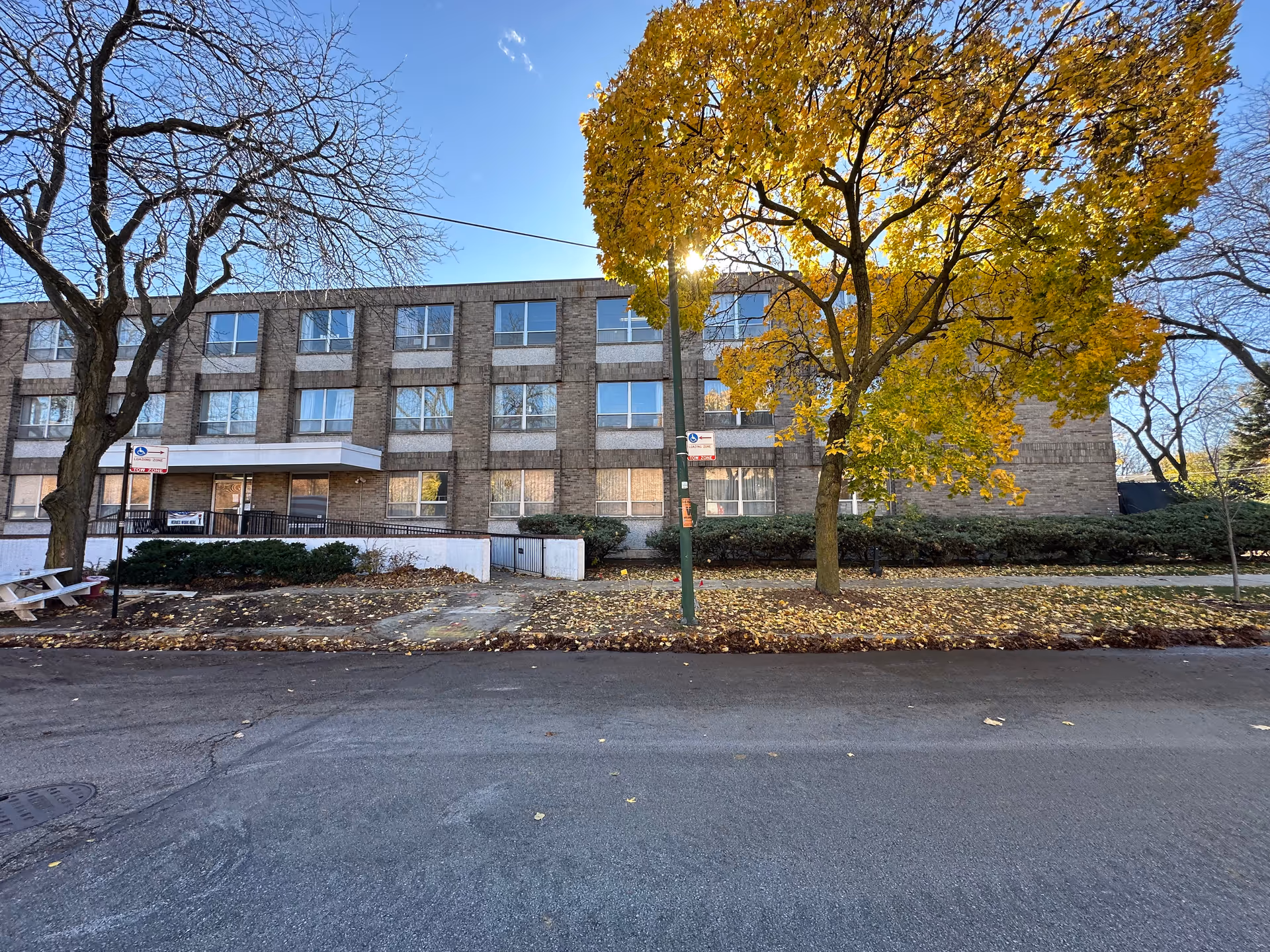 Three-story brick nursing home facade with a sidewalk and trees bearing yellow autumn leaves in front.