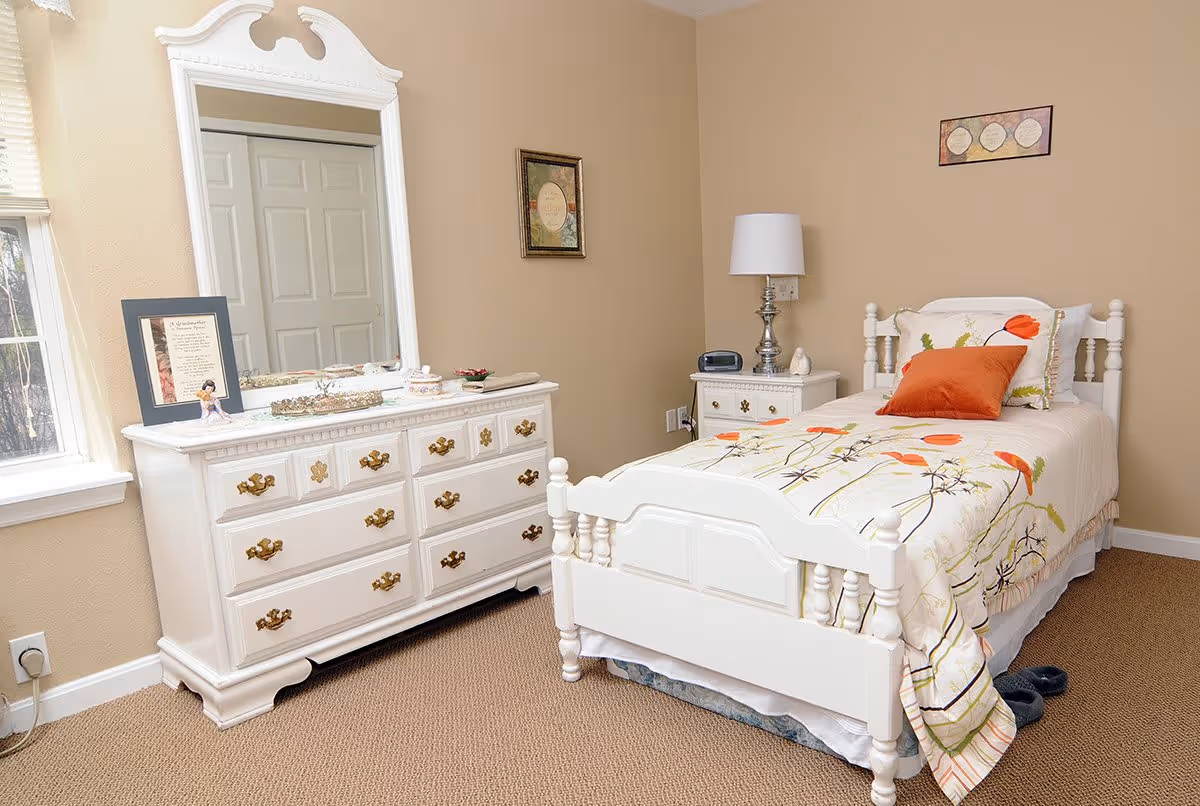 A cozy bedroom with a single white wooden bed featuring floral bedding and an orange pillow. Next to the bed is a white nightstand with a lamp and an alarm clock. Across from the bed is a white dresser with a large mirror, decorative items, and a framed picture. The walls are beige, and there is a window letting in natural light.