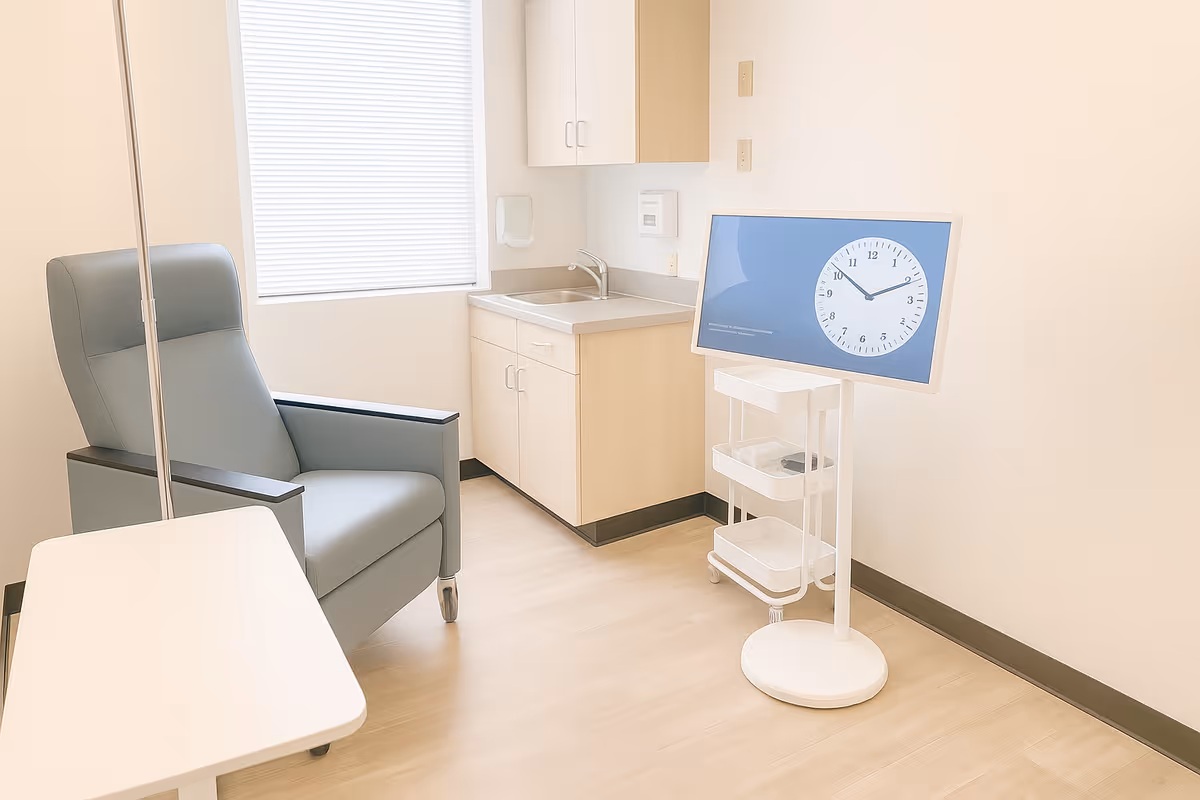 Clinic-style room featuring a gray patient chair, kitchenette, and a medical monitor on a stand.