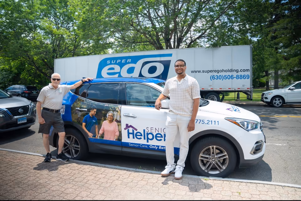 Two men pose beside a Senior Helpers promotional car in a parking lot.