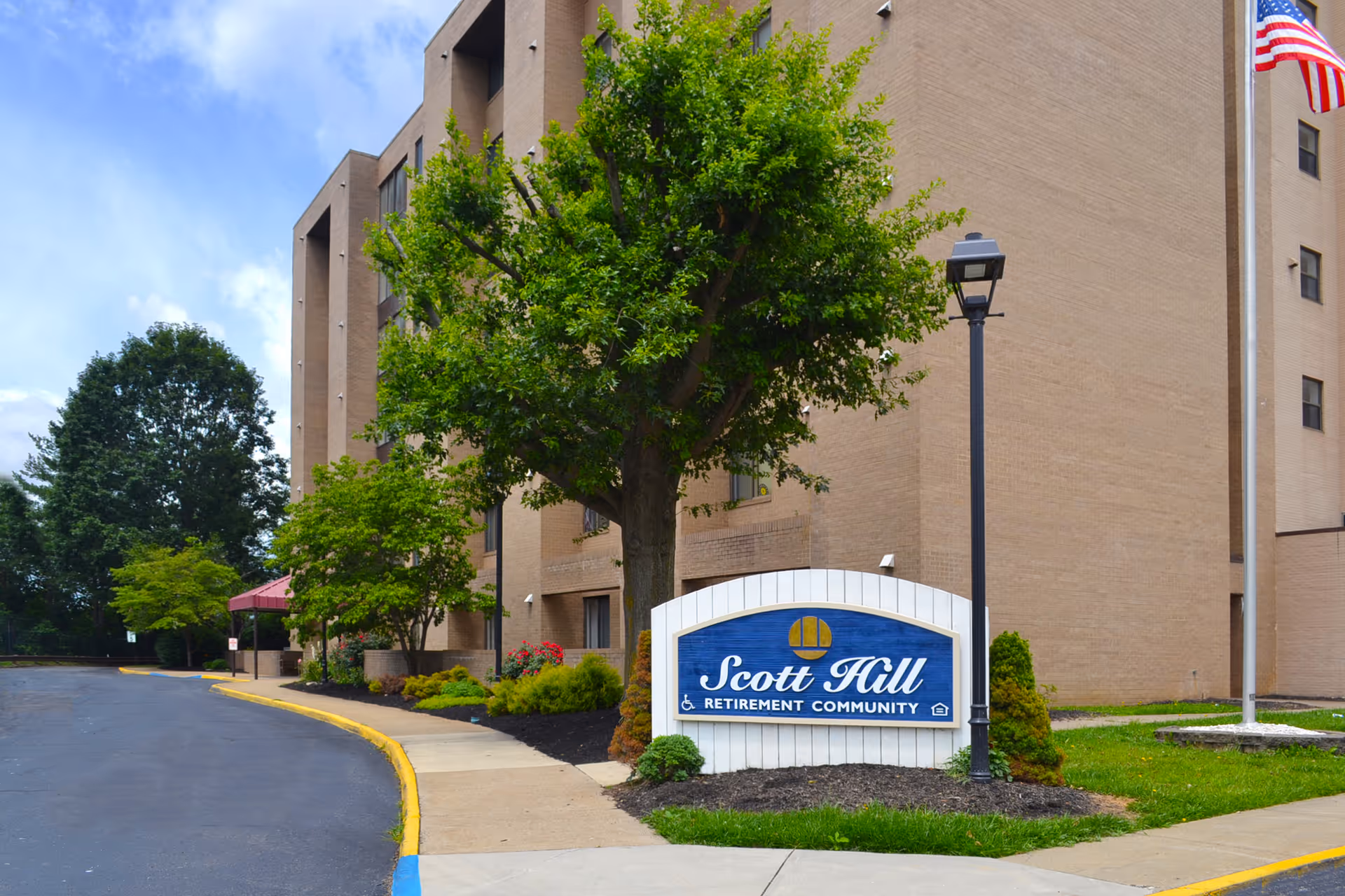Exterior front of Scott Hill Retirement Community showing a blue entrance sign, trees and landscaping, sidewalk, and a multi-story tan brick building.