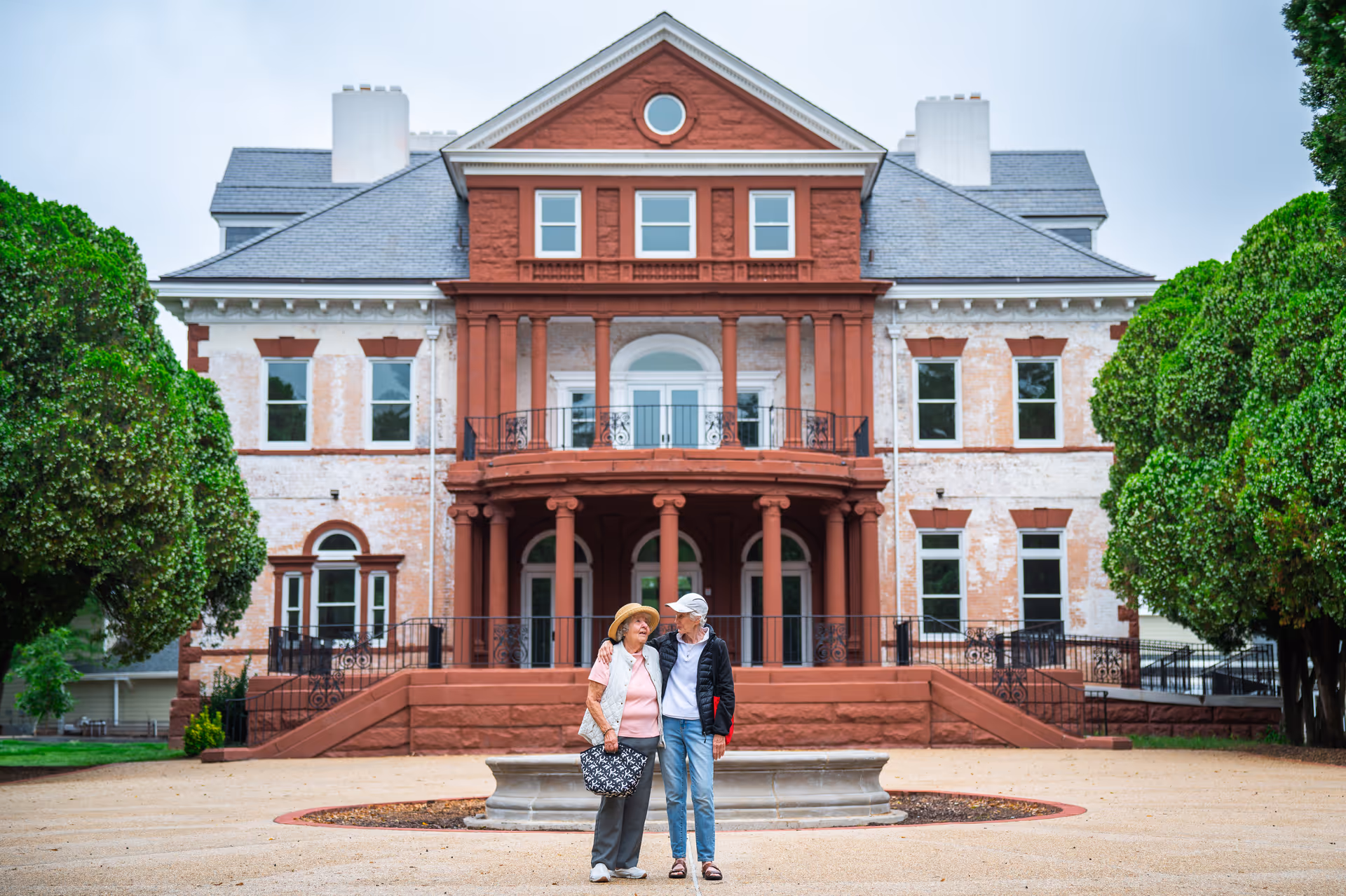 Historic red-brick manor-style building with two seniors posing outside.