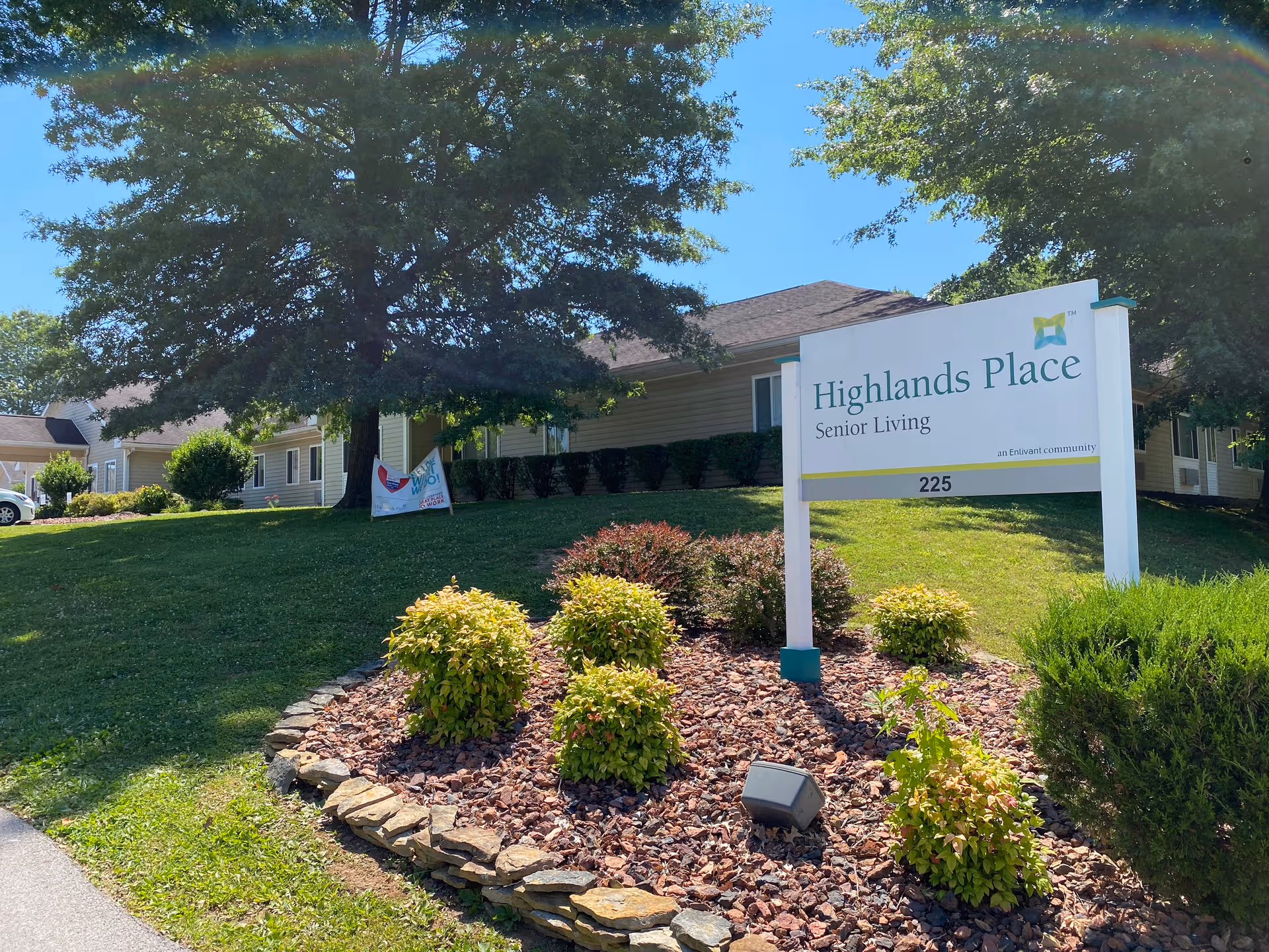 Exterior view of Highlands Place Senior Living with sign, trees, and landscaping.