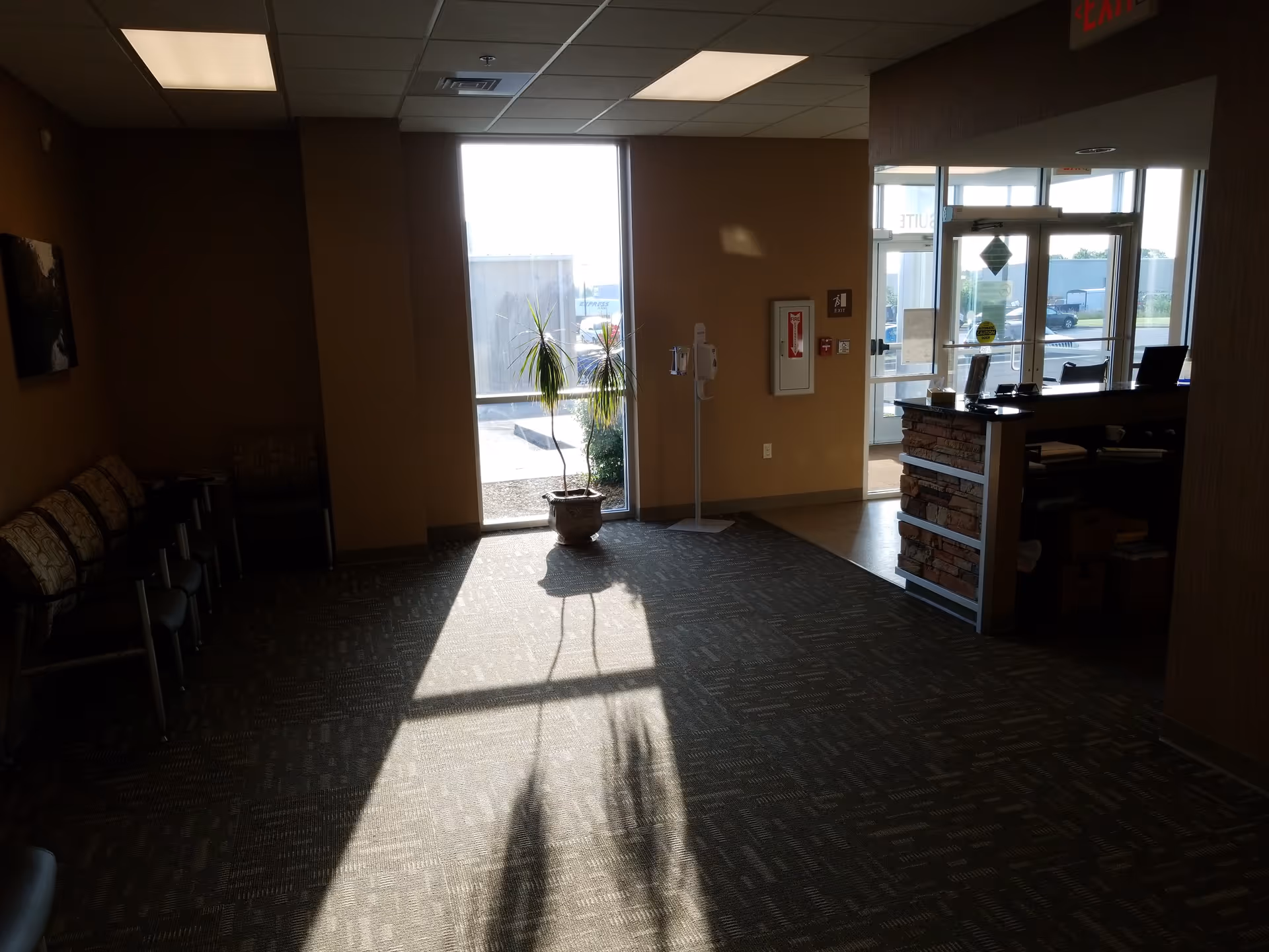Sunlit lobby with chairs along the left and a reception desk near glass doors.
