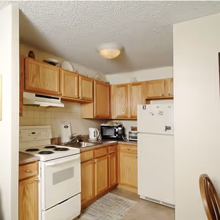 Compact L-shaped kitchen with wooden cabinets, stove, fridge, microwave, and sink.