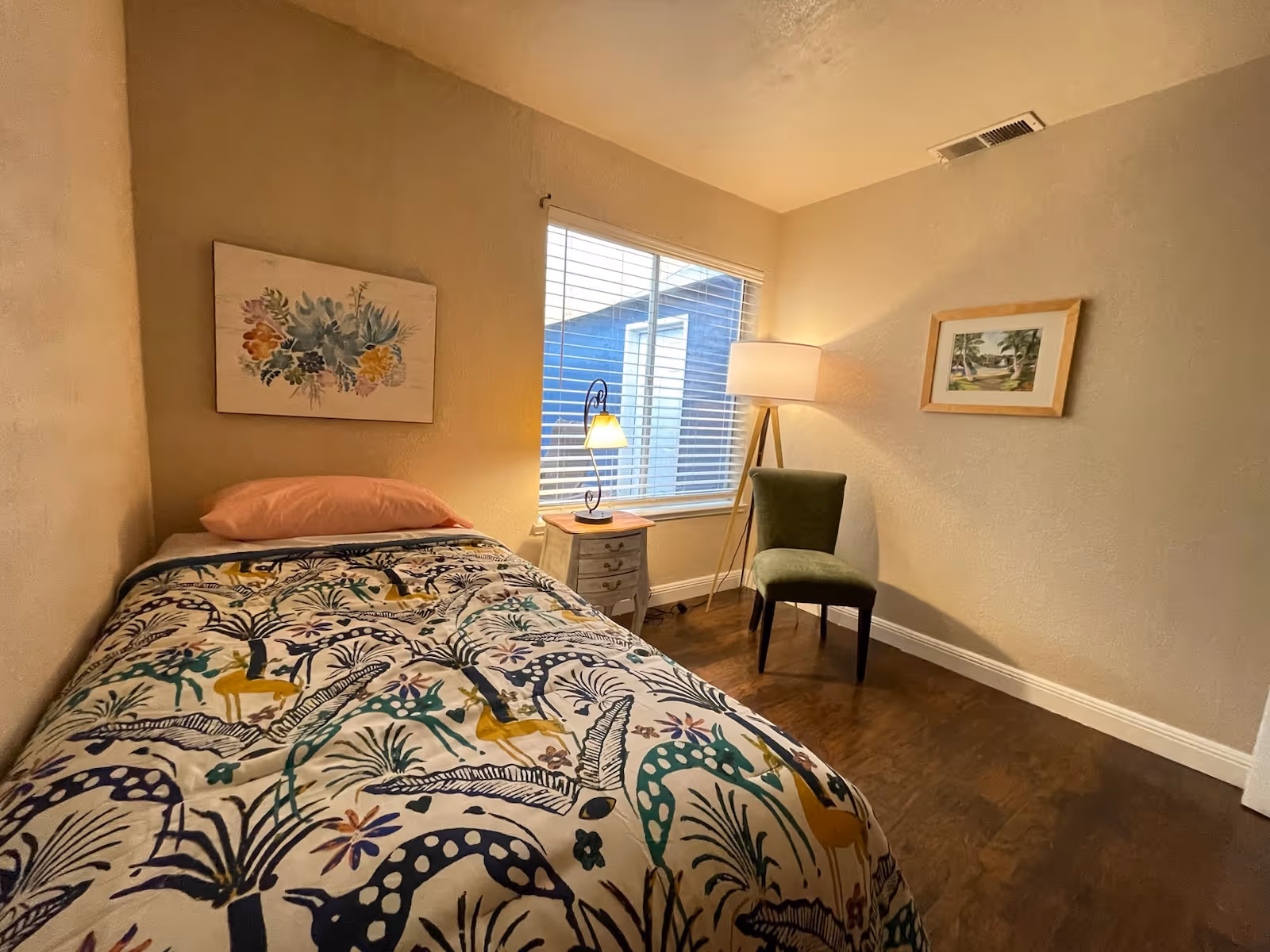 A small bedroom with a single bed covered in a colorful patterned blanket and a pink pillow. Next to the bed is a small nightstand with a lamp. There is a window with blinds letting in natural light. A green upholstered chair and a floor lamp are positioned in the corner. Two framed pictures hang on the beige walls. The floor is wooden.