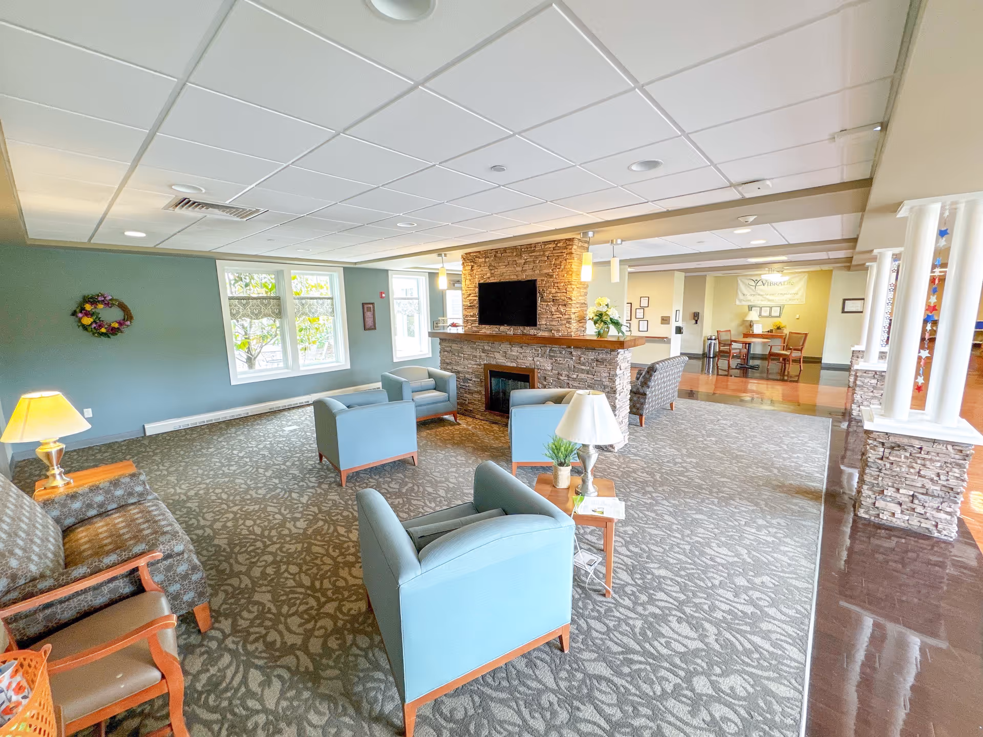 Spacious common lounge with blue armchairs arranged around a stone fireplace and mounted TV, lamps, and large windows.