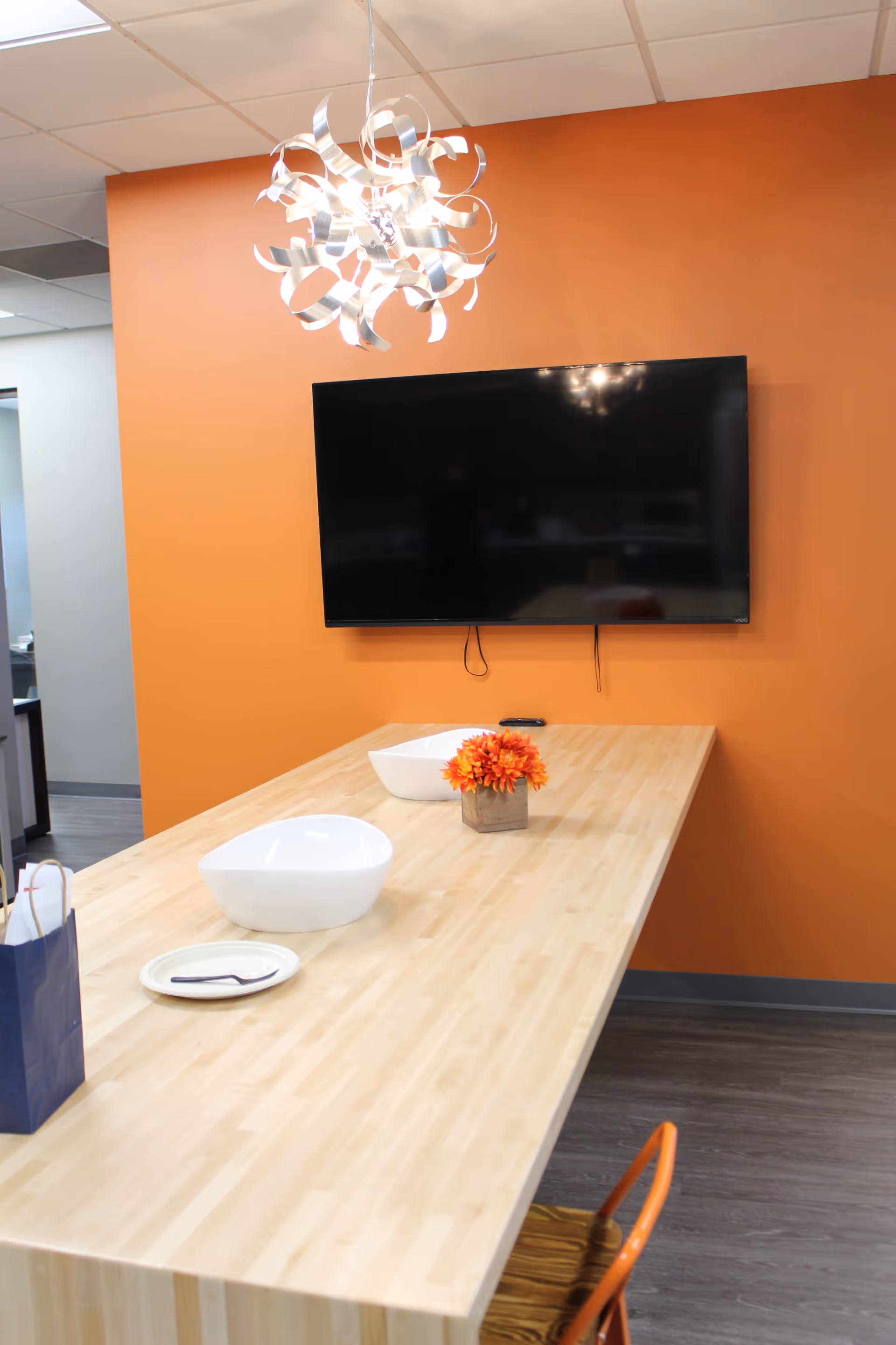 Orange-walled dining area with a long wooden table, bowls, flowers, and a wall-mounted TV.