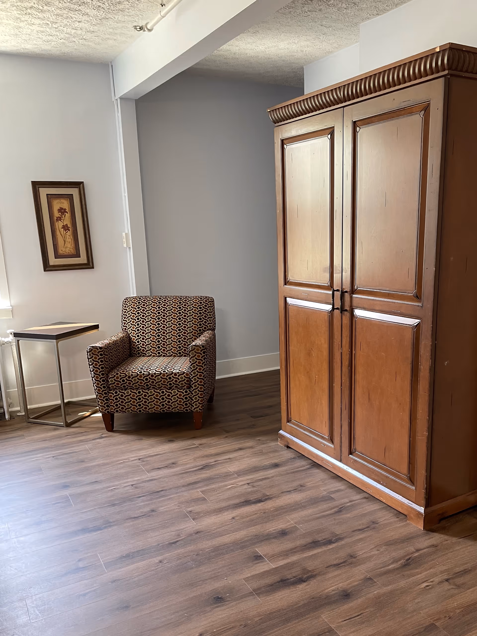 A patterned armchair beside a large wooden armoire in a simple living room.