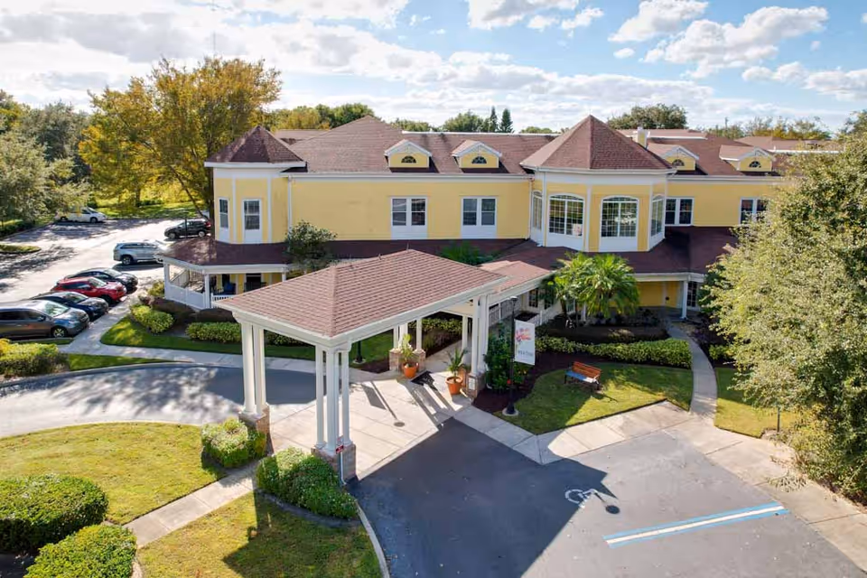 Exterior view of a two-story yellow senior living facility building with a covered entrance driveway, surrounded by trees and parked cars under a partly cloudy sky.