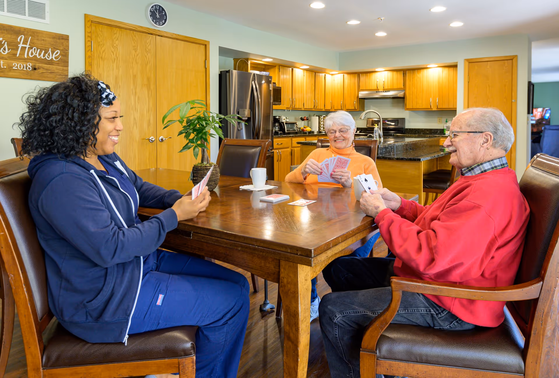 Three people sitting around a wooden dining table playing cards in a well-lit kitchen and dining area. The group includes an elderly woman in an orange sweater, an elderly man in a red sweater, and a younger woman in a navy blue outfit. The kitchen in the background has wooden cabinets, a refrigerator, and a countertop. A wooden sign on the wall reads 'Mimi's House Est. 2018'.