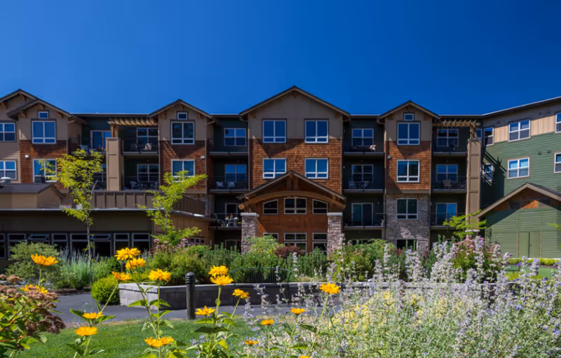 Front view of a multi-story senior living building with a landscaped flower garden in the foreground under a clear blue sky.
